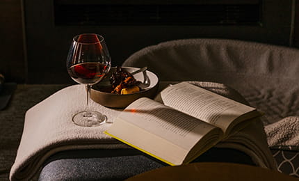 A glass of red wine, dessert, and opened book near the fireplace. 