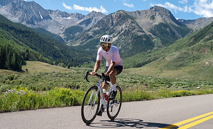A cyclist riding down the road on a summer day surrounded by mountains in the background.