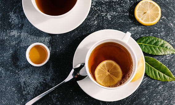 A cup of black tea on a stone surface surrounded by another lemon wheel, lemon leaves, and honey.