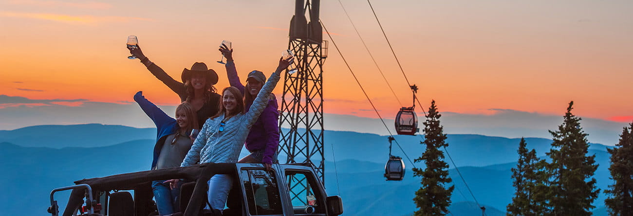 A group of people wave from the open top of a Jeep during an Off-Road Tour, with the Aspen Mountain Gondola and sunset in the background.