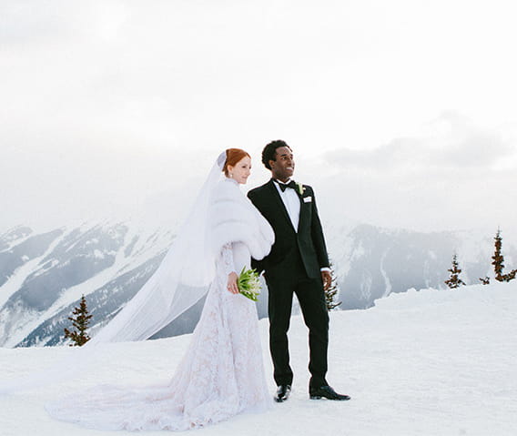 A bride in her wedding dress and groom in his tuxedo on top of the snow capped Aspen Mountain in the winter. 