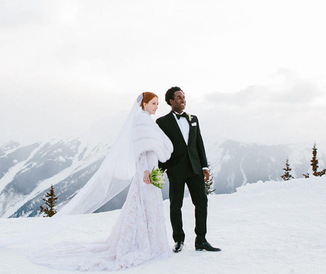 A bride in her wedding dress and groom in his tuxedo on top of the snow capped Aspen Mountain in the winter. 