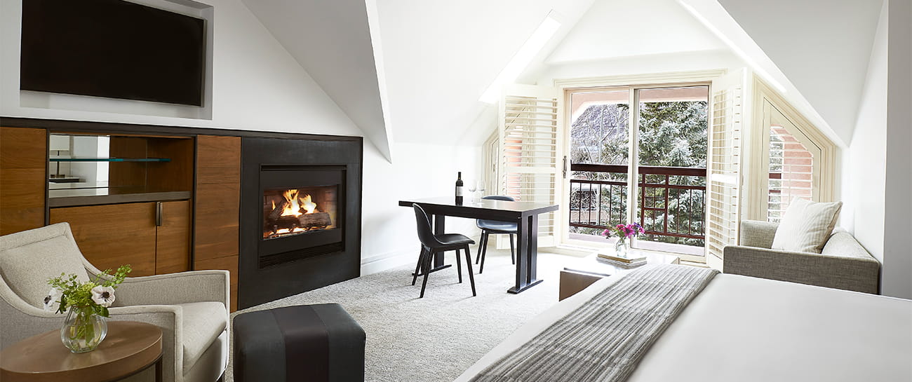 Interior of a spacious room at The Little Nell with a king size bed, lit fire place, and snowy trees outside the window.
