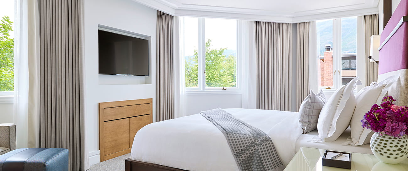 View of a large premium hotel room, with queen bed, wall mounted tv, and tree filled windows.
