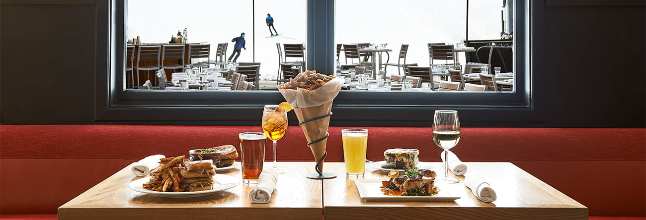 A table at Ajax Tavern with classic dishes and beverages, and skiers through the winter coming down Aspen Mountain.