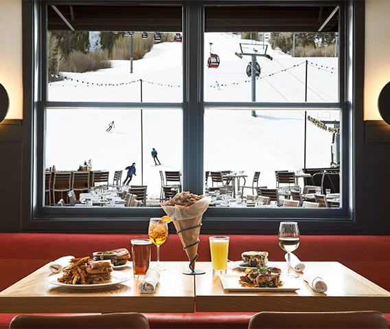 A table at Ajax Tavern with classic dishes and beverages, and skiers through the winter coming down Aspen Mountain.