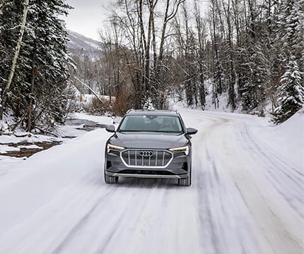 An Audi driving down a snowy road near an icy creek and the woods.